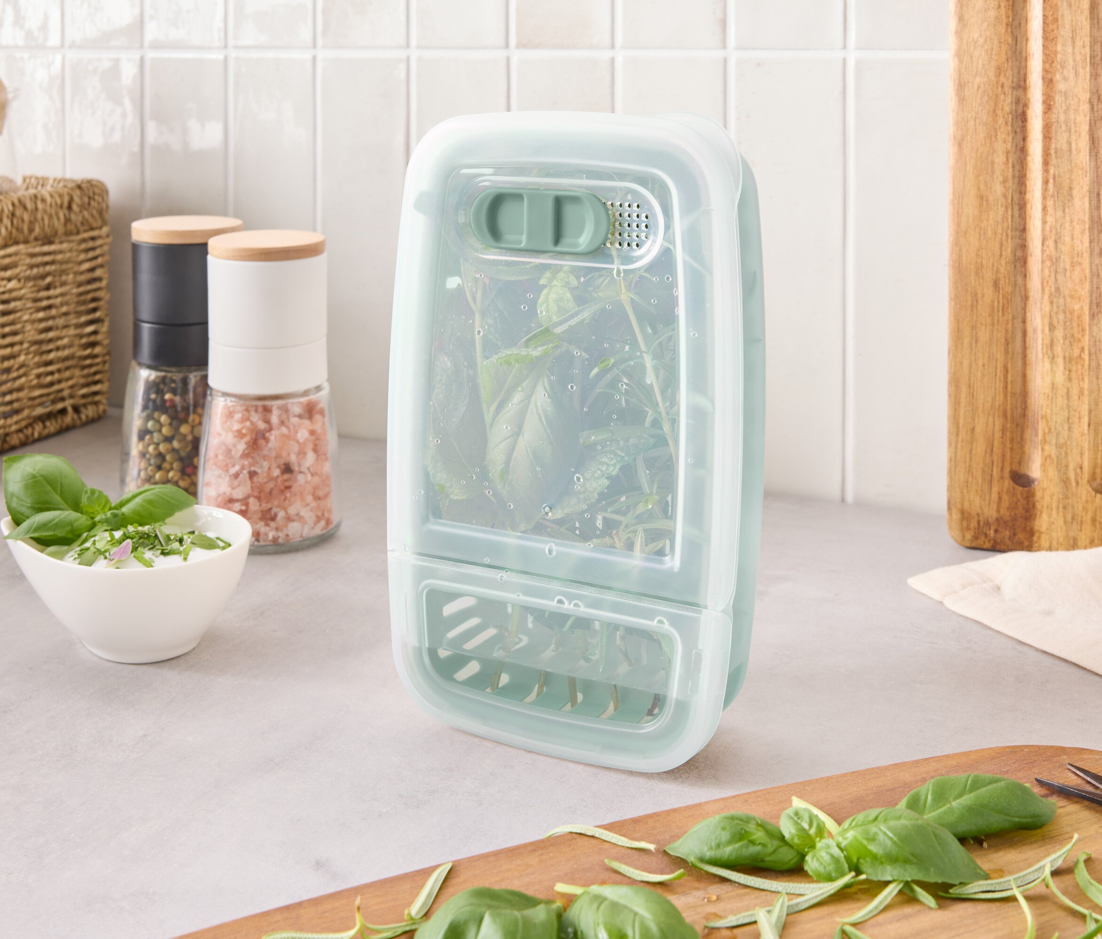 Ein Kräuteraufbewahrungsbehälter mit Kräutern steht auf einer Küchentheke neben Zutaten und einem Schneidebrett.