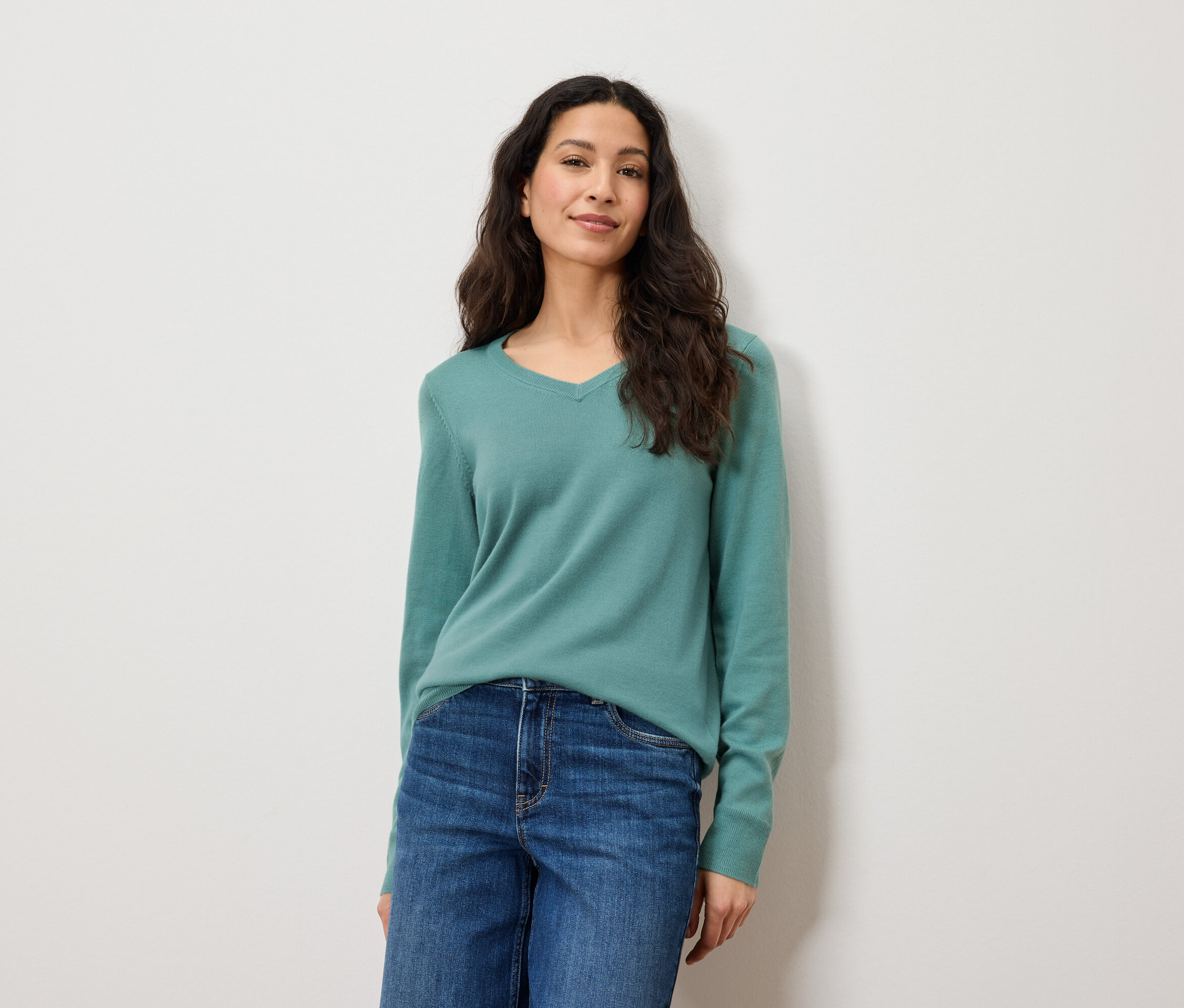 Eine Frau mit langen braunen Haaren posiert vor einem weißen Hintergrund. Sie trägt blaue Jeans und einen grünen Pullover.