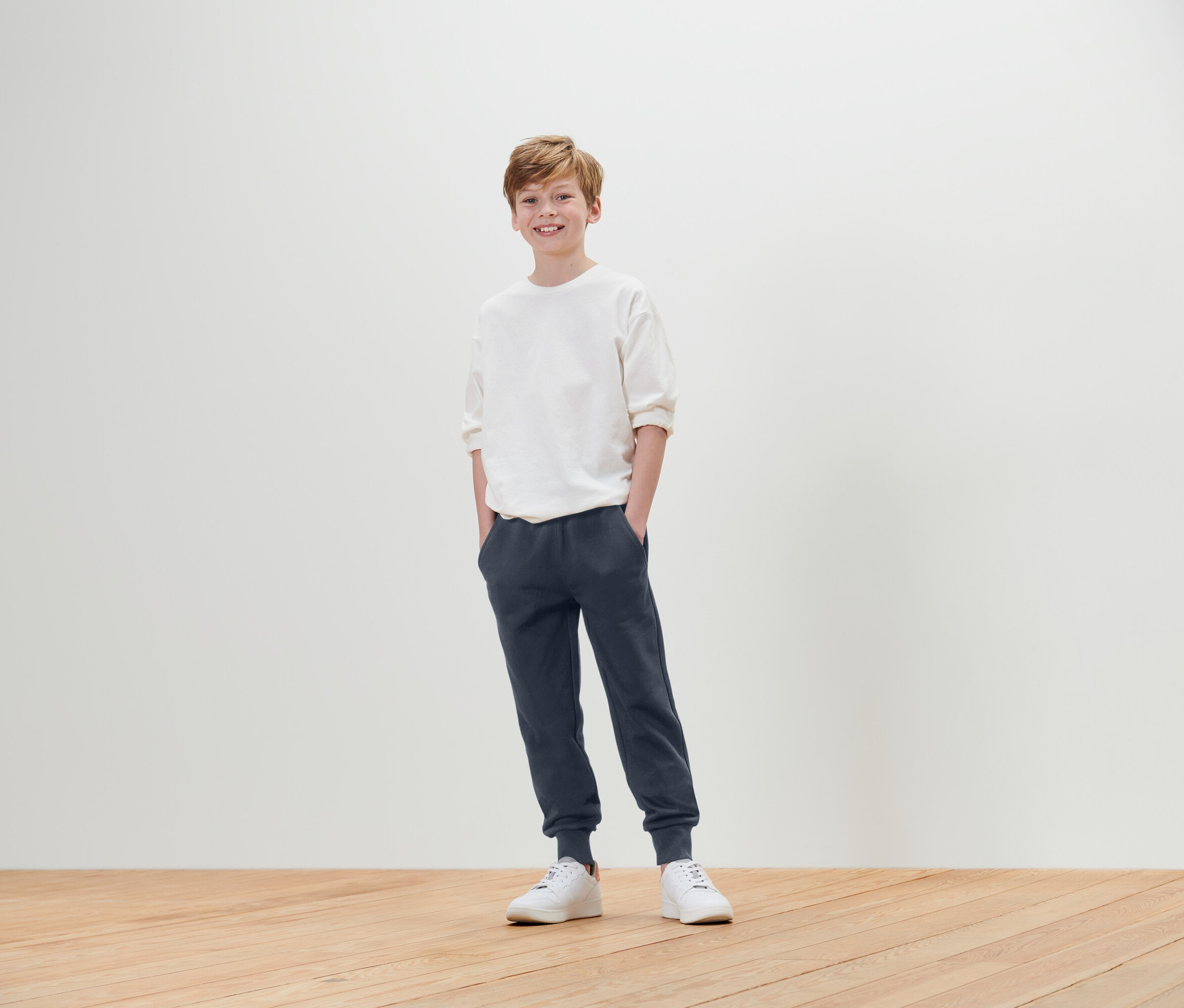 Ein Junge steht mit den Händen in den Hosentaschen und trägt ein weißes T-Shirt, eine blaue Jogginghose und weiße Turnschuhe.
