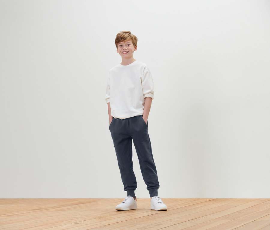 Ein Junge steht mit den Händen in den Hosentaschen und trägt ein weißes T-Shirt, eine blaue Jogginghose und weiße Turnschuhe.