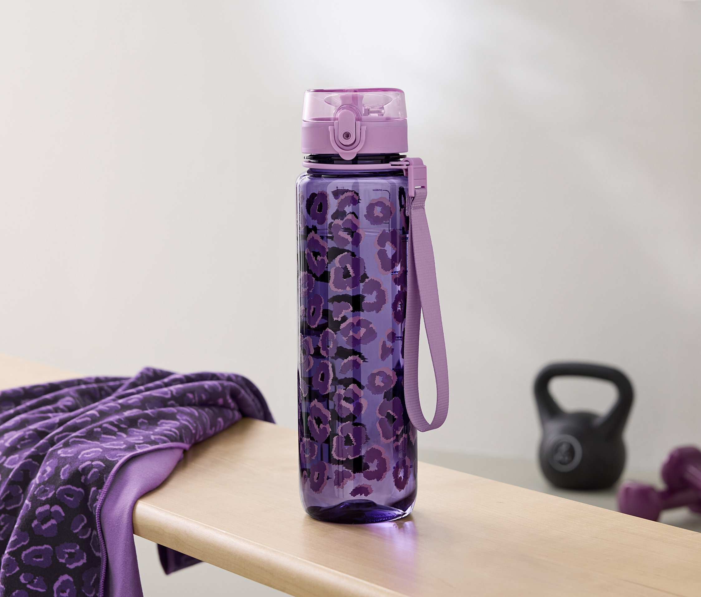 Eine lila Wasserflasche mit Muster liegt neben einem Handtuch und Hanteln auf einer Bank.