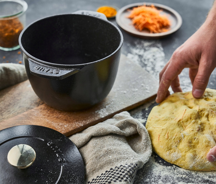 Hände formen Teig neben einer schwarzen 16 cm Staub Reis-Cocotte auf einem Holzbrett.