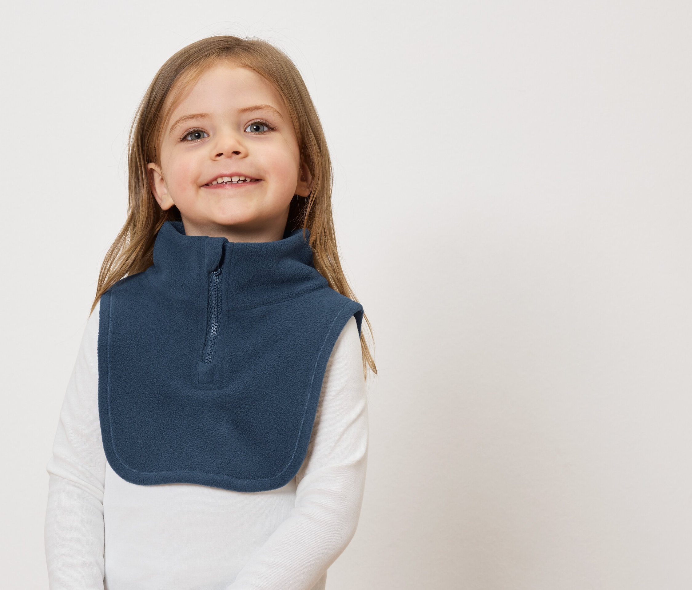 Lächelndes Mädchen mit braunen Haaren und einem blauen Fleece-Kragenwärmer vor einem weißen Hintergrund.