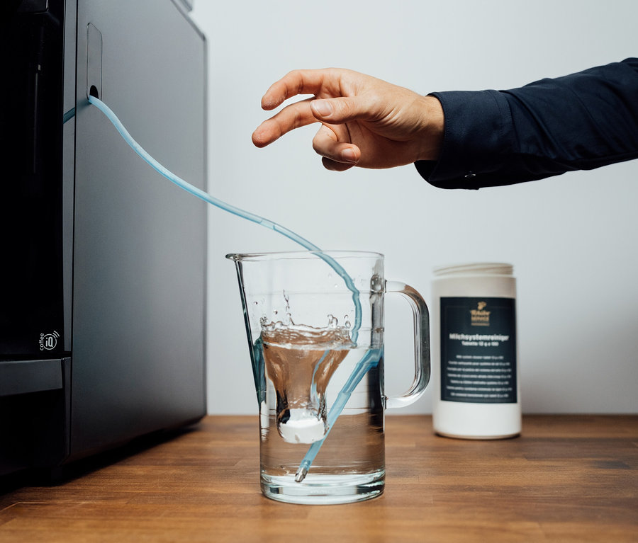 Eine Hand zeigt auf einen Schlauch, der von einer Kaffeemaschine in einen Krug mit Wasser führt. Daneben steht eine Packung Milchsystemreiniger Tablette 12g x100.