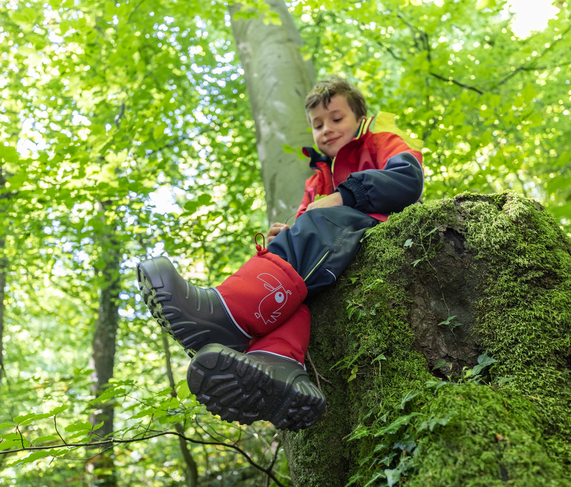 Ein Junge sitzt auf einem mit Moos bewachsenen Baumstumpf im Wald und trägt rote Gummistiefel.