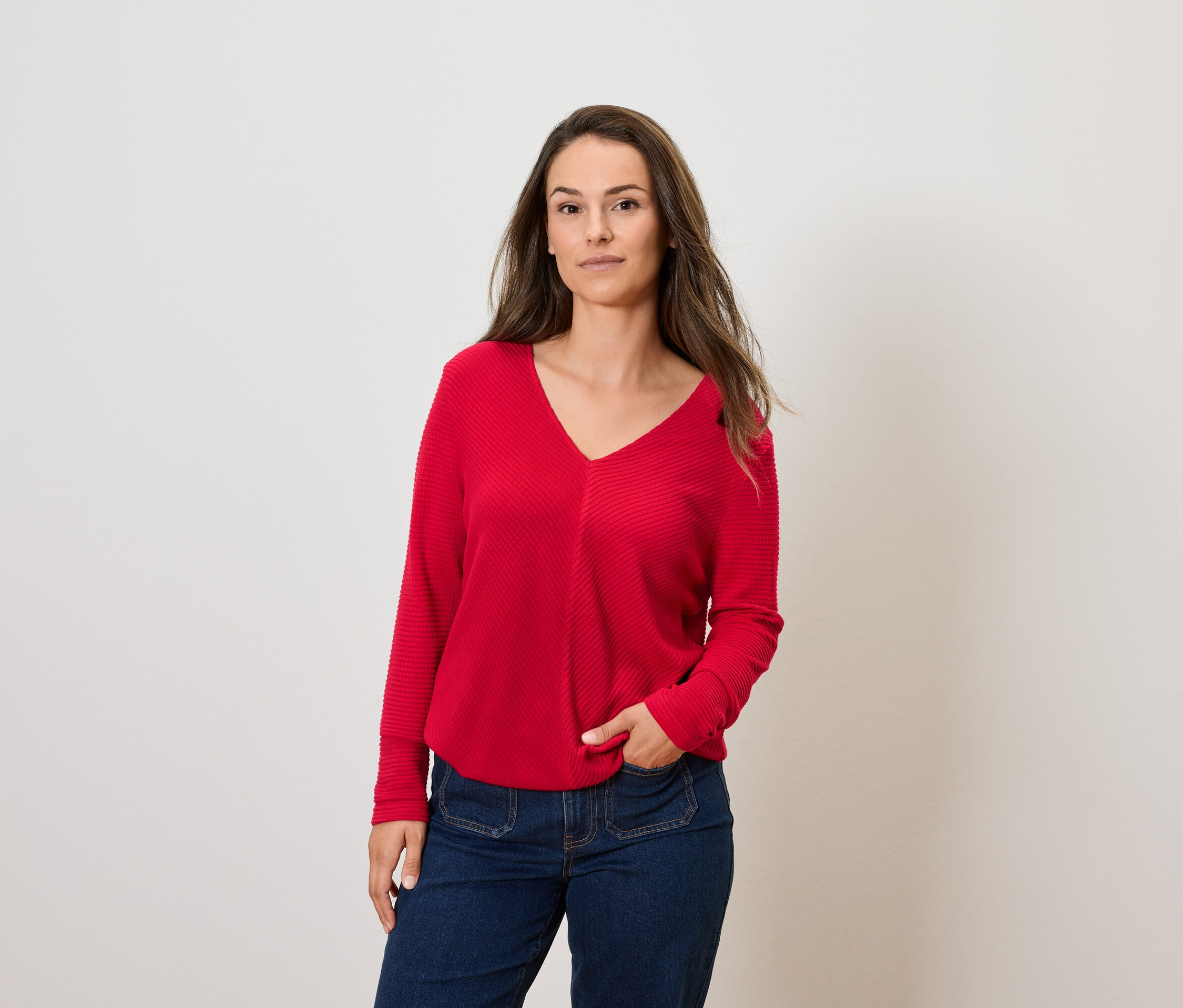 Frau mit braunem Haar posiert in rotem Langarmpullover und blauen Jeans.