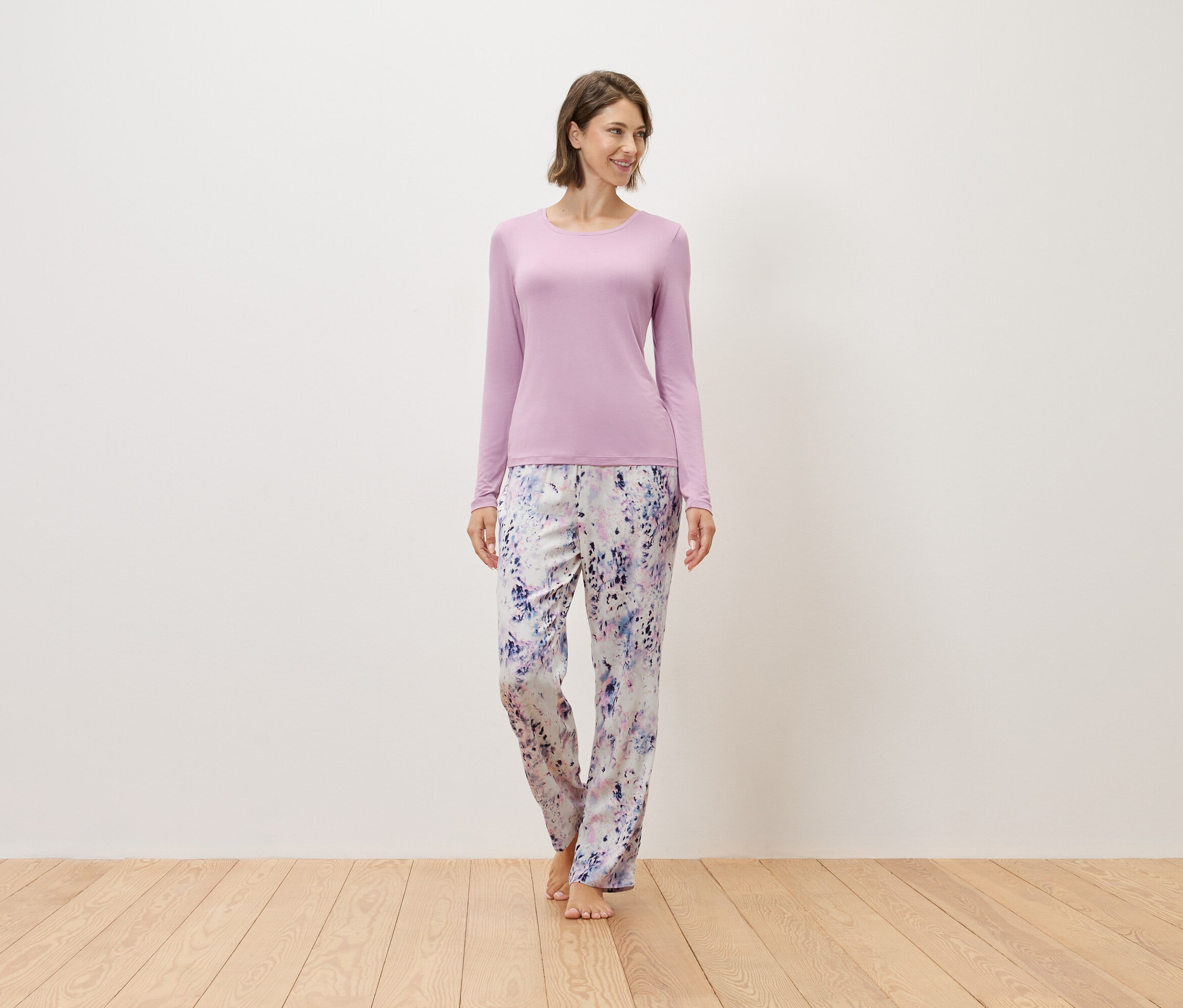 Frau steht barfuß auf einem Holzboden, trägt einen lila Pullover und eine bedruckte Hose.