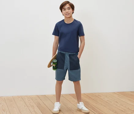 Junge hält ein Skateboard und trägt Kinder-Bermudashorts.