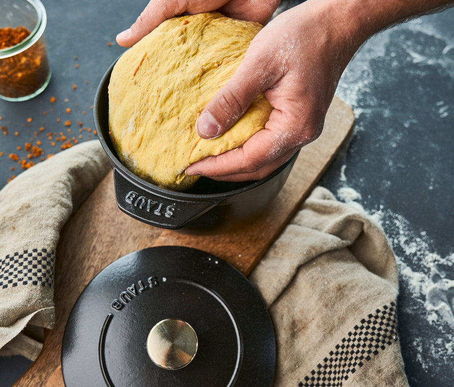 Hände legen gelben Teig in eine schwarze Staub Reis-Cocotte, 16 cm. Der Deckel liegt daneben auf einem Brettchen.