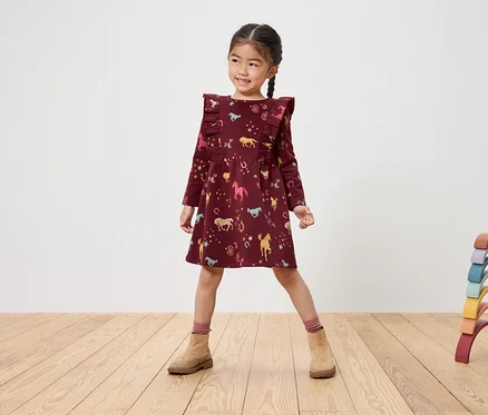 Lächelndes Mädchen in bordeauxfarbenem Kleid mit Pferdeaufdruck und braunen Stiefeln steht auf einem Holzboden neben einem regenbogenfarbenen Holzspielzeug.