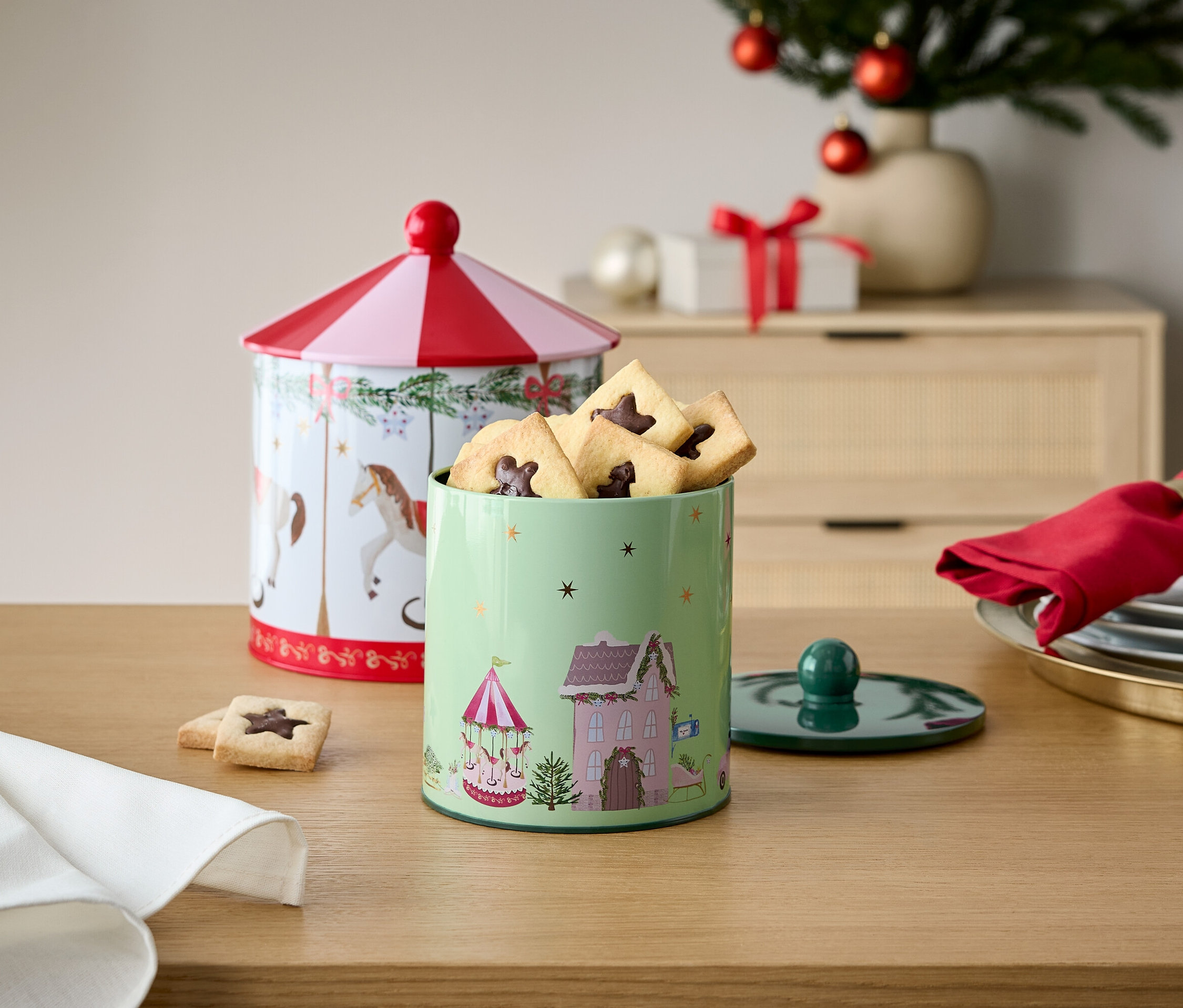 Zwei dekorierte Keksdosen aus Blech auf einem Holztisch mit Weihnachtsschmuck im Hintergrund.