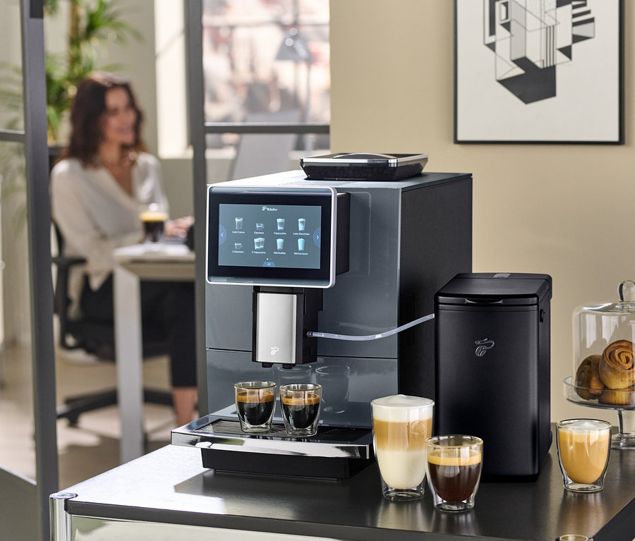 Ein Kaffeevollautomat bereitet Kaffee in Gläsern auf einem Tisch zu. Im Hintergrund sitzt eine Frau an einem Tisch.