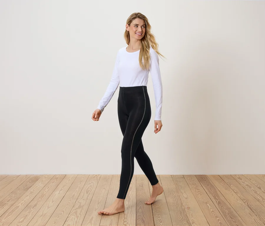 Eine Frau geht barfuß auf einem Holzboden und trägt schwarze Leggings und ein weißes Langarmshirt.