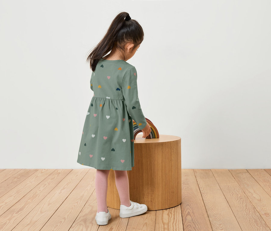Mädchen in grünem Kleid mit Herzen steht an einem Holztisch mit einem Regenbogen.