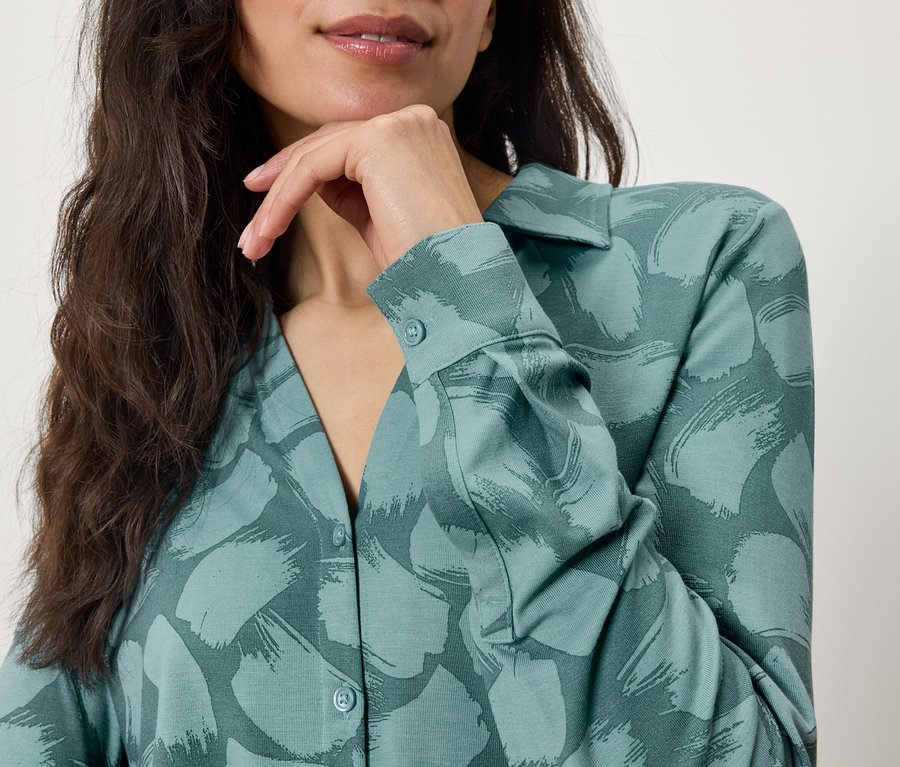 Oberkörper einer Frau mit braunen Haaren und einer grün gemusterten Bluse, die Hand am Kinn.