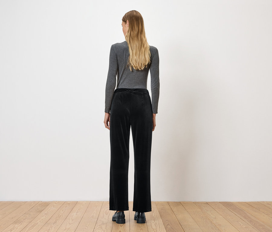 Frau steht mit dem Rücken zur Kamera und trägt ein graues Langarmshirt und eine schwarze Samthose.