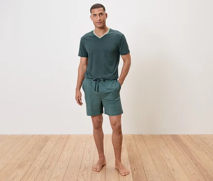 Mann steht barfuß auf Holzboden und trägt einen grünen Schlafanzug mit kurzen Ärmeln und Shorts.