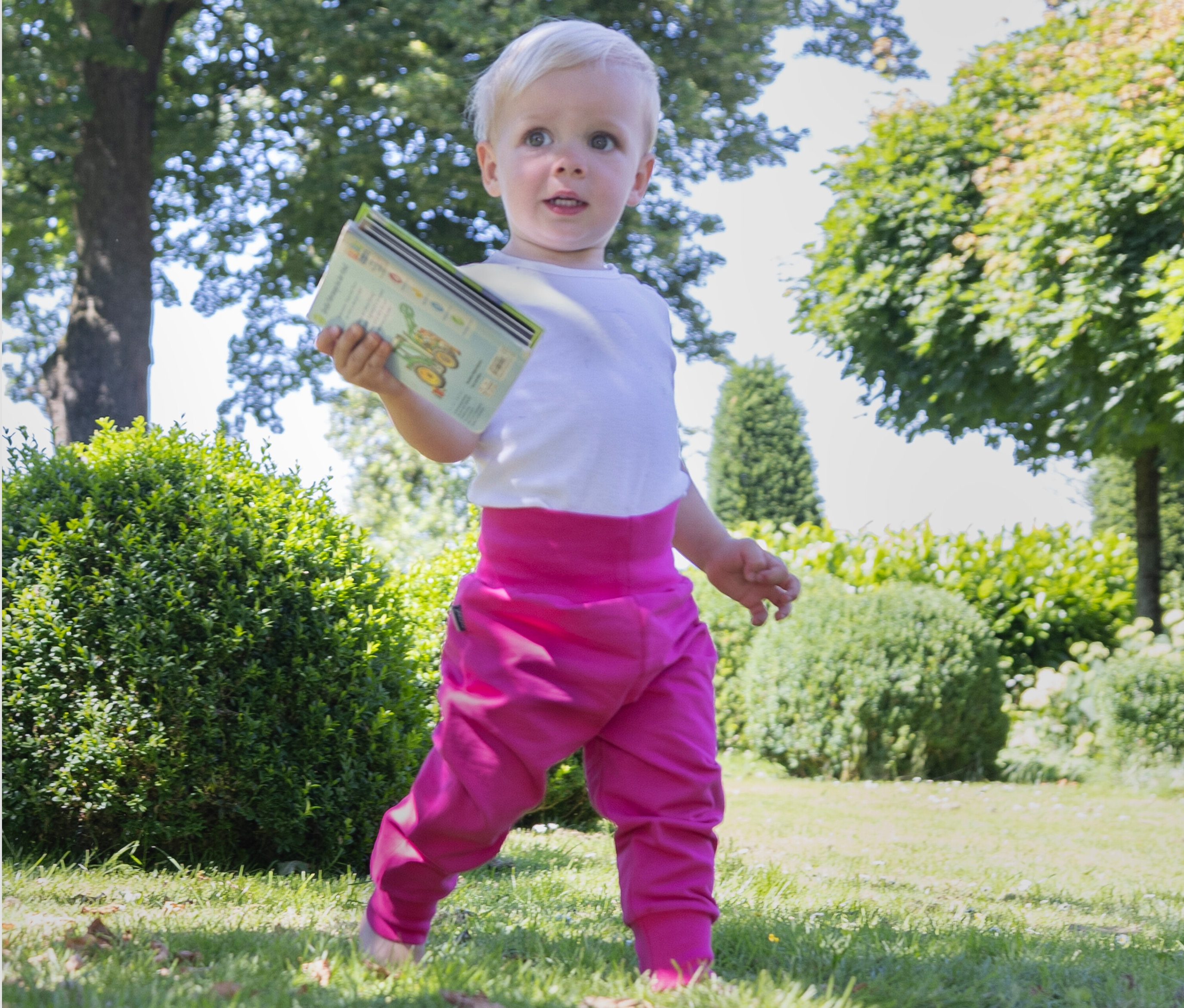 Kleinkind mit blonden Haaren hält ein Buch und steht auf einer Wiese. Es trägt ein weißes T-Shirt und eine fuchsiafarbene Jako-O Baby Wohlfühlhose.