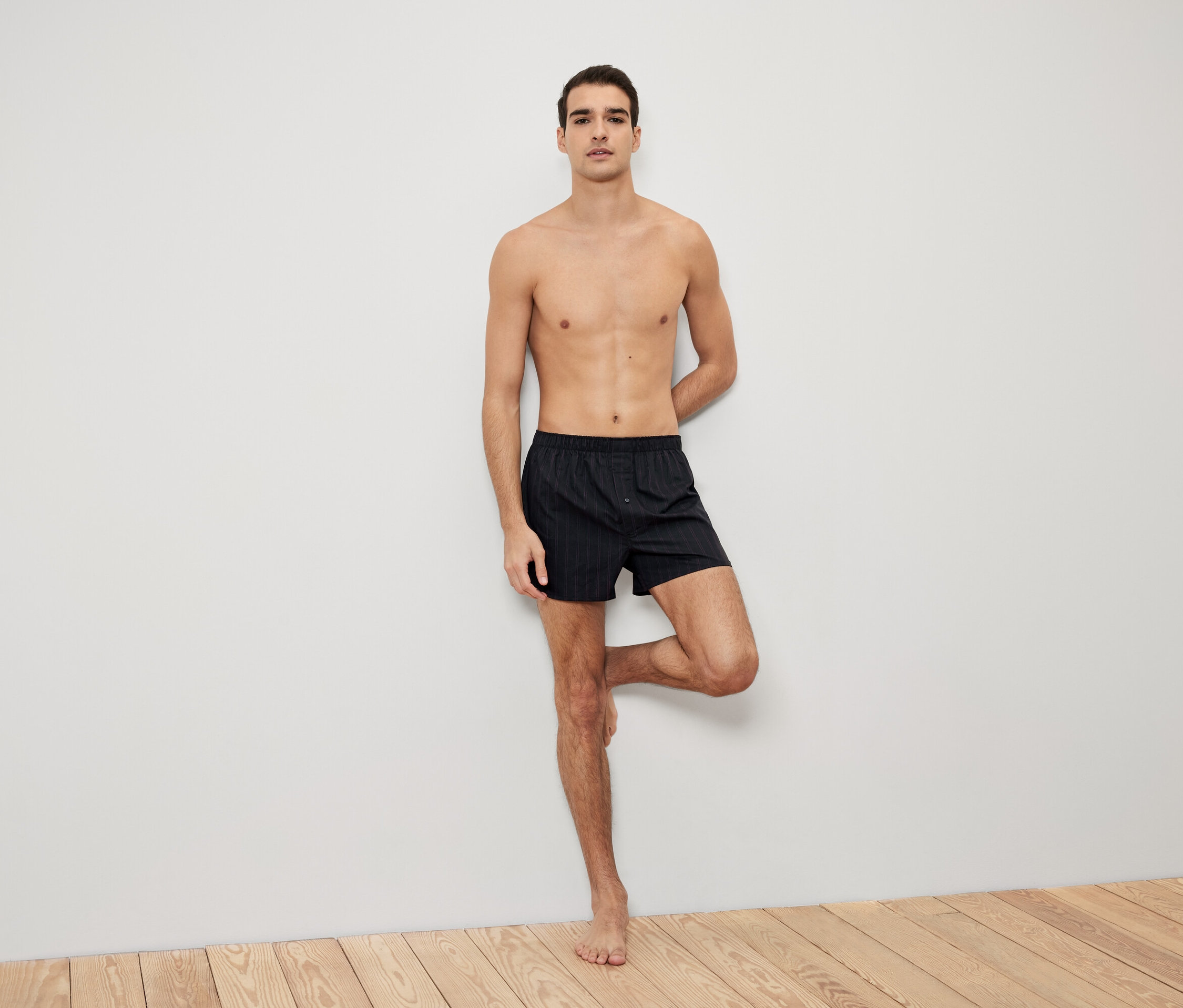 Mann posiert an eine Wand gelehnt in schwarzen gestreiften Boxershorts.