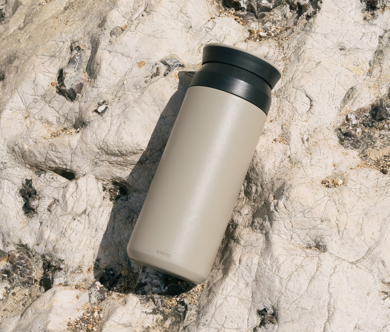 Eine hellbeige Kinto-Flasche mit einem schwarzen Deckel liegt auf einem hellen, zerklüfteten Felsen.