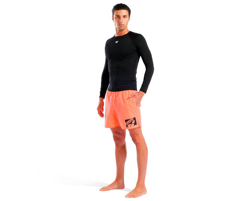 Mann steht barfuß in Badeshorts und einem schwarzen Langarmshirt vor einem weißen Hintergrund.