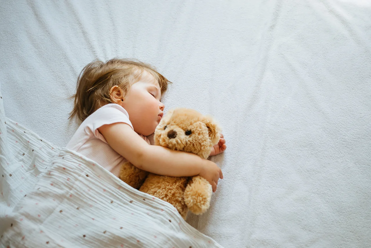 Ein schlafendes Kind umarmt einen Teddybären unter einer Decke auf einem weißen Bettlaken.