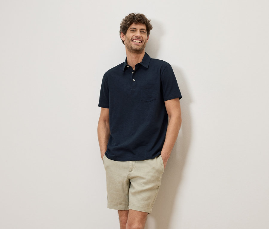 Ein lächelnder Mann mit lockigem Haar steht vor einer Wand und trägt ein marineblaues Poloshirt und Shorts.