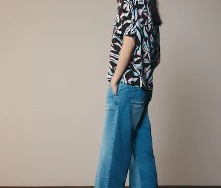 Eine Frau mit langen dunklen Haaren steht mit den Händen in den Taschen und blickt über ihre Schulter. Sie trägt eine gemusterte CECIL Kurzarmbluse in Schwarz mit weißen, blauen und rosa Mustern, weite Jeans und helle Sandalen.