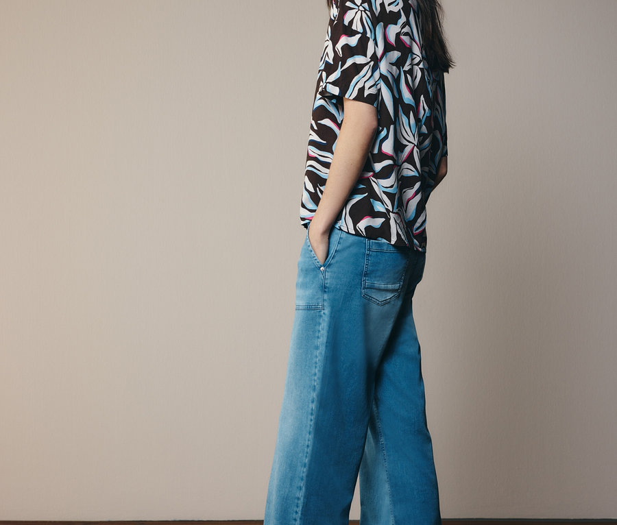 Frau mit gemustertem Oberteil und ausgestellter Jeanshose, die zur Seite schaut.