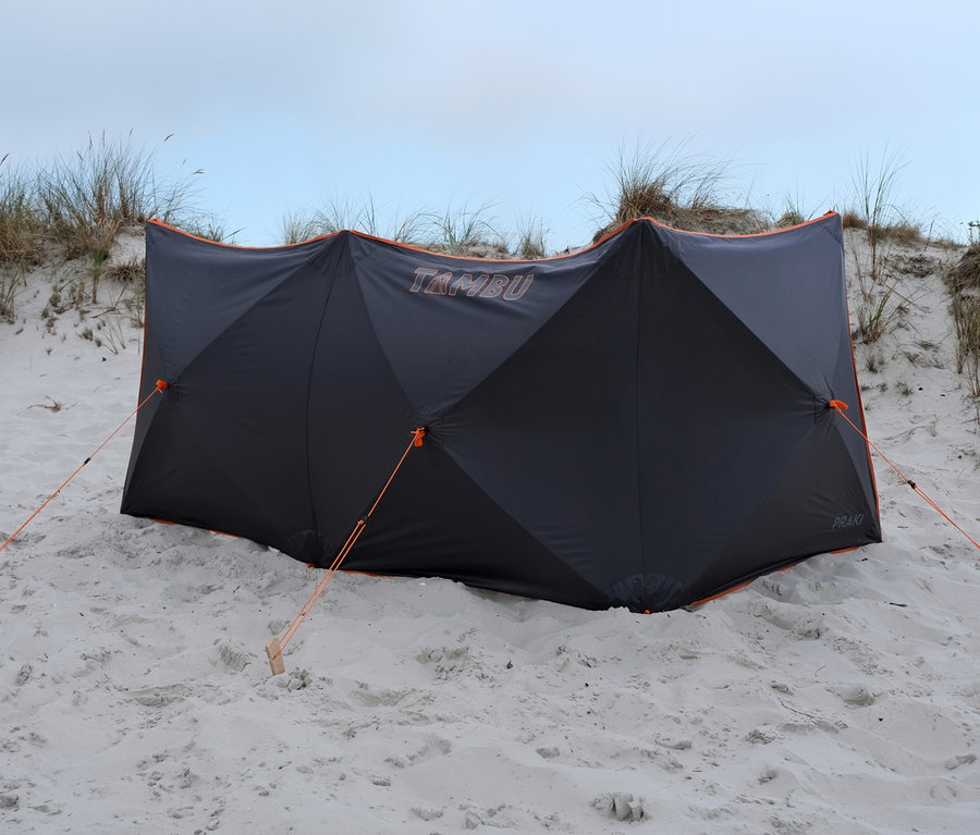 Ein grauer Windschutz mit orangefarbenen Akzenten steht am Strand, befestigt mit orangefarbenen Seilen und Holzpflöcken.