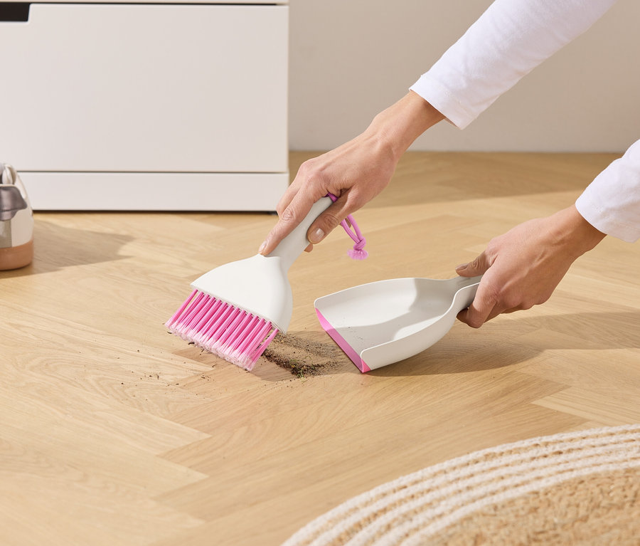 Eine Person kehrt mit einer weißen Kehrschaufel und einer pinkfarbenen Bürste Schmutz auf einem Holzboden auf.