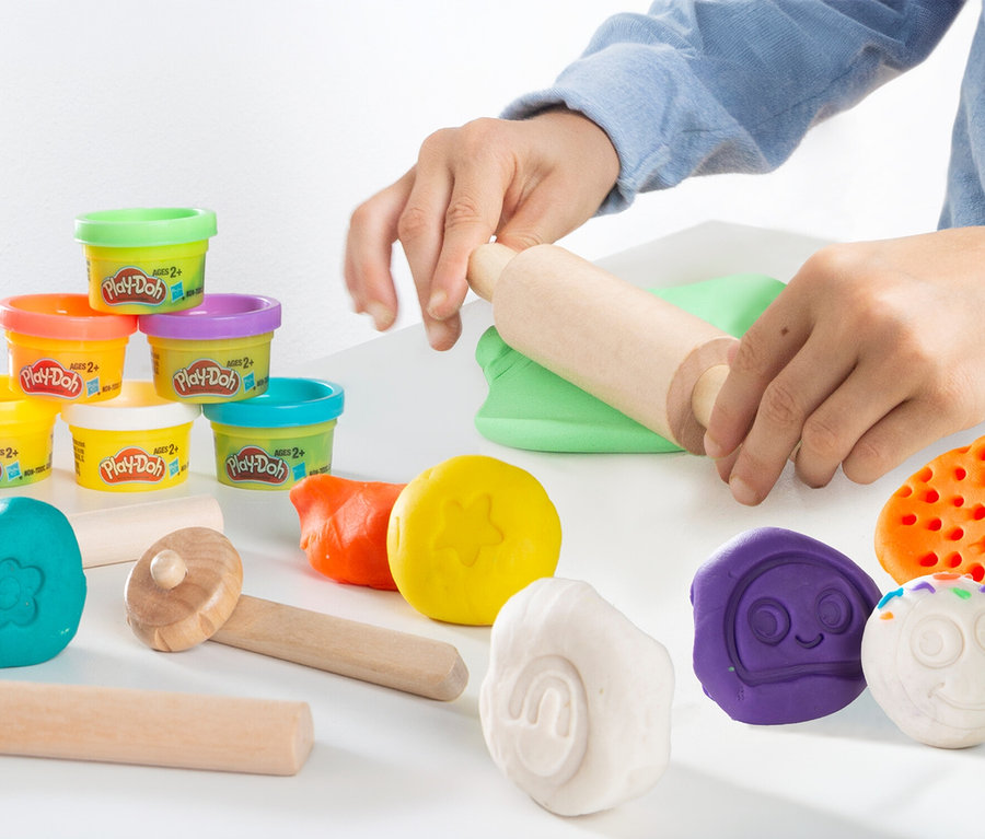 Eine Person rollt grüne Play-Doh mit einem Nudelholz aus, neben bunten Play-Doh-Dosen und Formen.