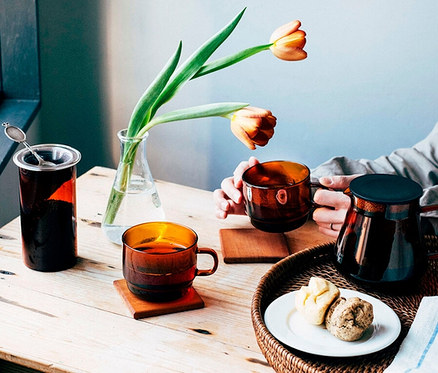 Eine Person hält eine amberfarbene Tasse aus dem HARIO »Colors« Tee- und Kaffee-Set. Auf dem Tisch steht das restliche Set mit Gebäck.