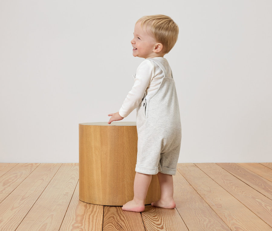 Kleiner Junge in hellgrauem Overall steht neben einem runden Holzhocker.