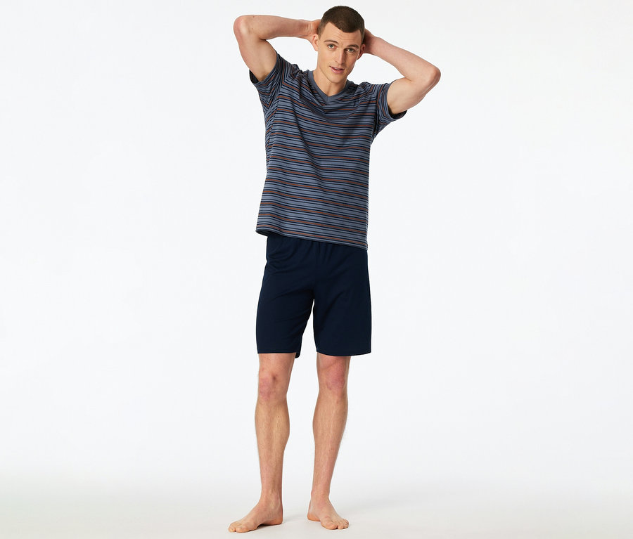 Mann steht barfuß mit erhobenen Händen im gestreiften T-Shirt und blauen Shorts vor einem weißen Hintergrund.