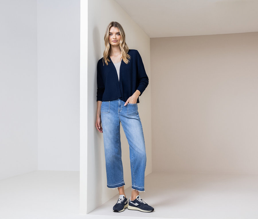 Frau lehnt an einer Wand, trägt eine marineblaue Strickjacke, blaue Jeans und marineblaue Schuhe.