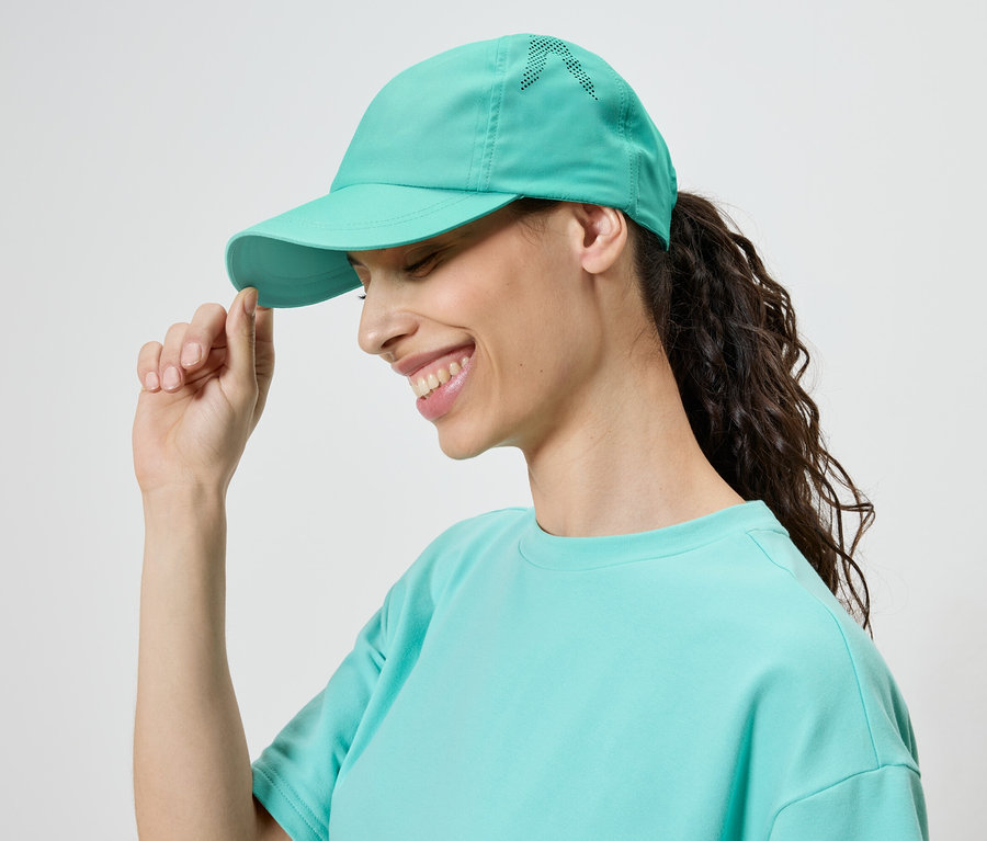 Lächelnde Frau mit einem türkisfarbenen Cap und T-Shirt, die den Schirm der Kappe berührt.