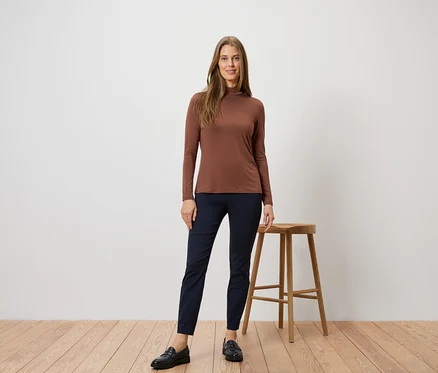 Frau in braunem Rollkragenshirt und blauen Hosen posiert neben einem Hocker auf einem Holzboden.
