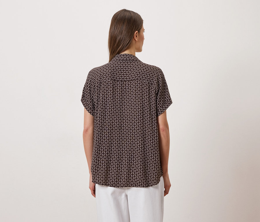Rückansicht einer Frau mit braunen Haaren in einer braunen, kurzärmeligen Bluse mit geometrischem Muster und weißer Hose.