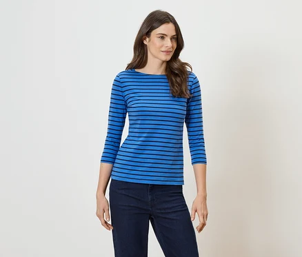 Eine Frau mit langen, gewellten, braunen Haaren trägt ein blaues, horizontal gestreiftes Oberteil und eine dunkle Jeans.