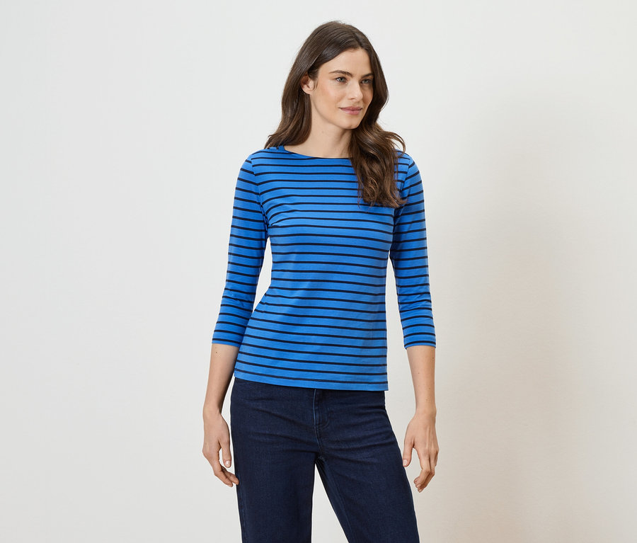Eine Frau mit langen, gewellten, braunen Haaren trägt ein blaues, horizontal gestreiftes Oberteil und eine dunkle Jeans.