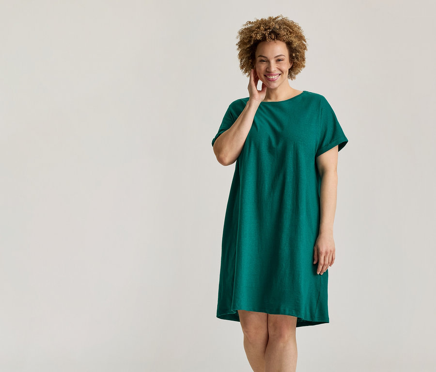 Lächelnde Frau mit lockigem Haar in einem grünen Kurzarmkleid, die ihr Gesicht berührt.