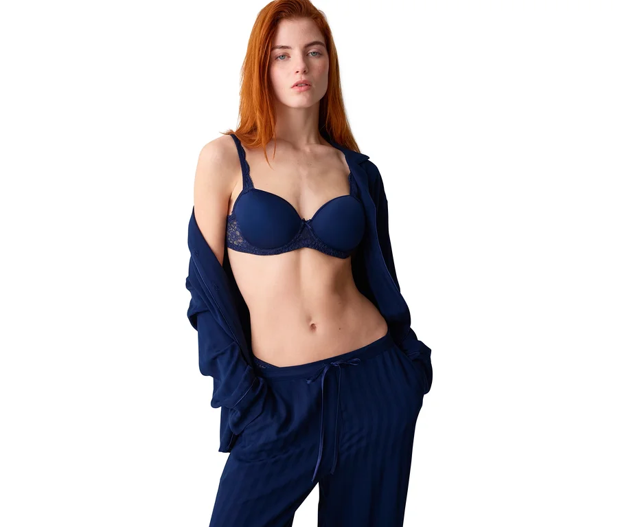 Eine rothaarige Frau steht vor weißem Hintergrund und trägt einen marineblauen BH sowie passende Pyjamahosen. Ein marineblaues Hemd ist über ihre Schultern gelegt.