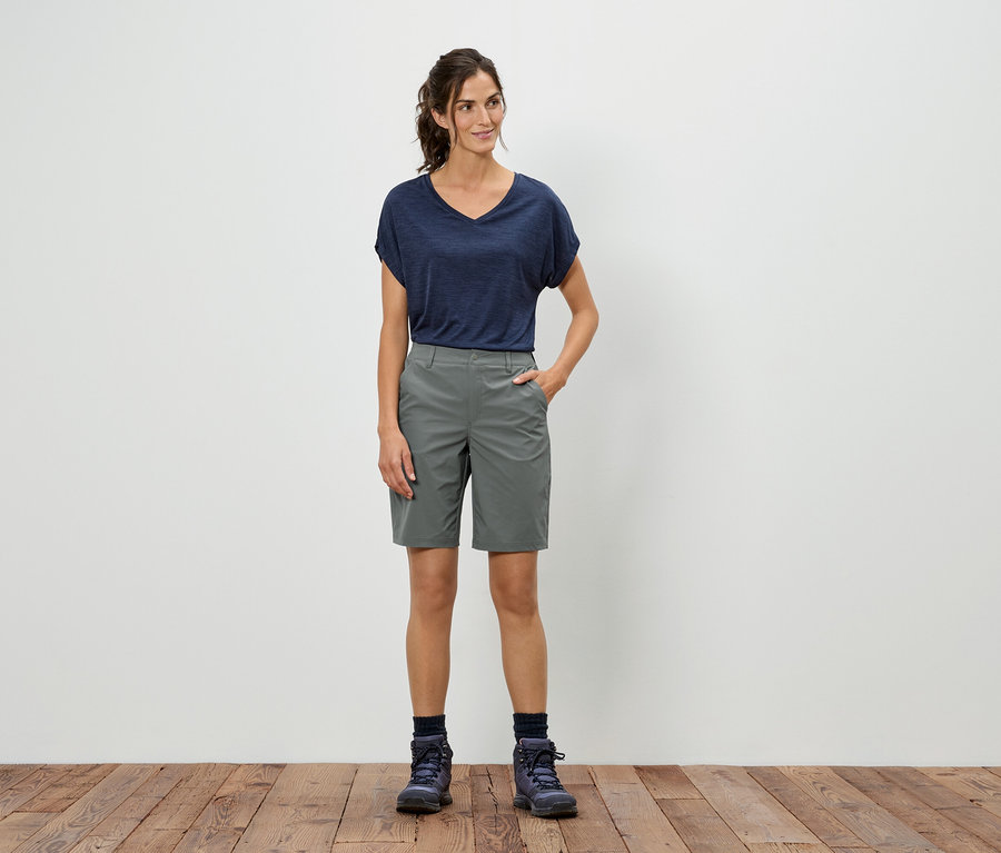 Eine Frau steht vor einer weißen Wand und trägt ein blaues T-Shirt, graue Shorts und Wanderschuhe.