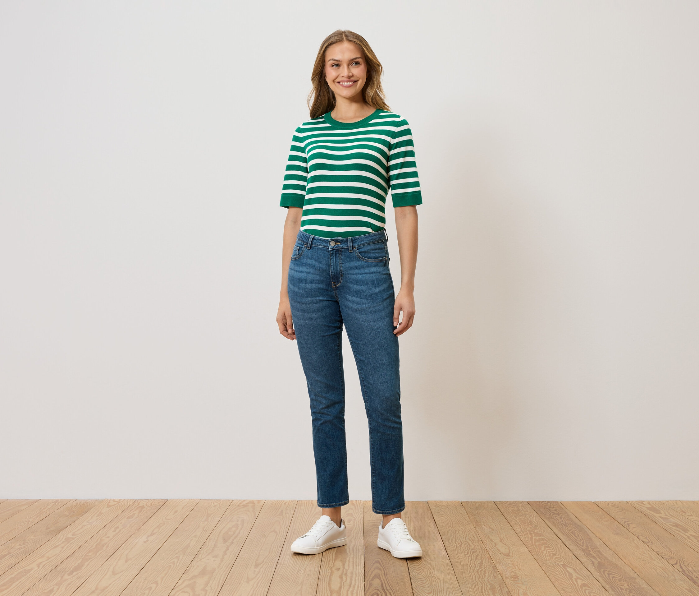 Frau steht und trägt ein grün-weiß gestreiftes Oberteil, blaue Jeans und weiße Turnschuhe.