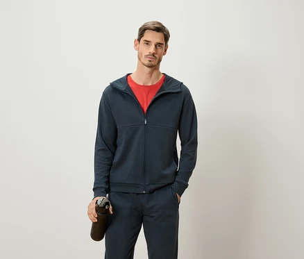 Mann mit rotem T-Shirt, dunkelblauer Trainingsjacke und Trainingshose hält eine dunkle Wasserflasche vor einem weißen Hintergrund.