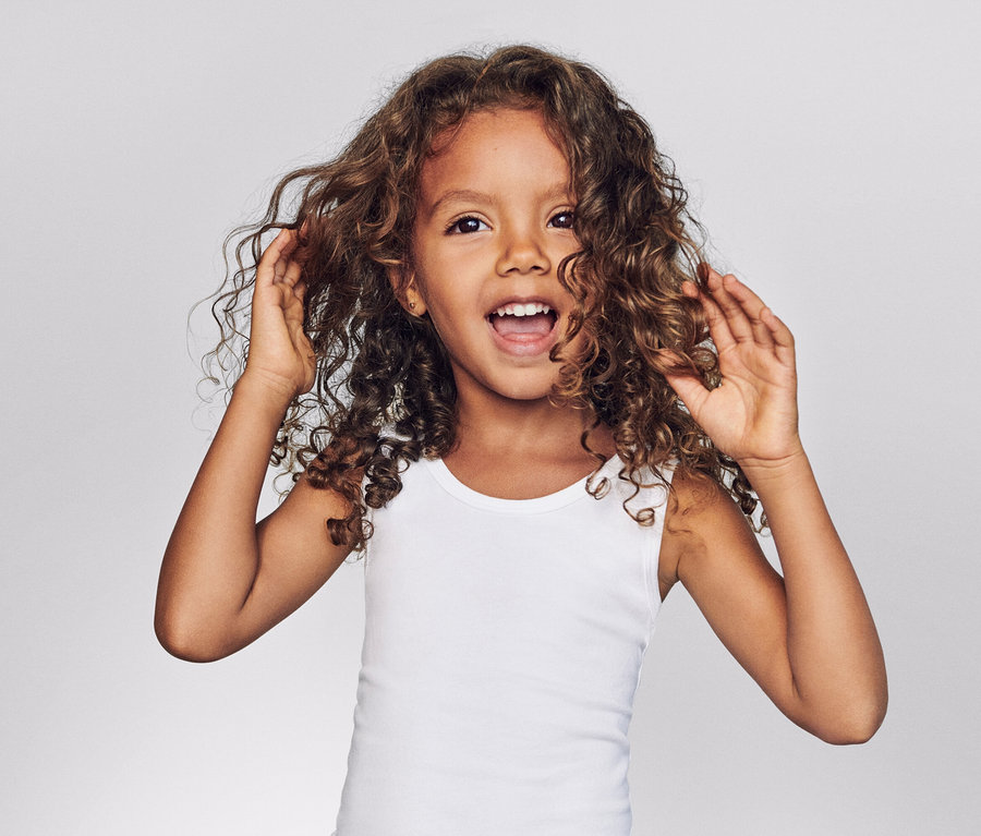 Mädchen mit lockigem Haar und weißem Tanktop lacht und berührt ihr Haar vor einem grauen Hintergrund.