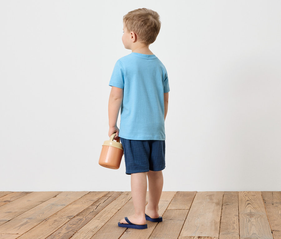 Ein Junge von hinten trägt ein blaues T-Shirt, Shorts und blaue Flip-Flops und hält einen braunen Behälter in der Hand.