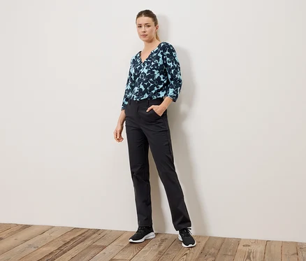 Frau steht mit Hand in der Hosentasche, trägt eine blau gemusterte Bluse und schwarze Hose.