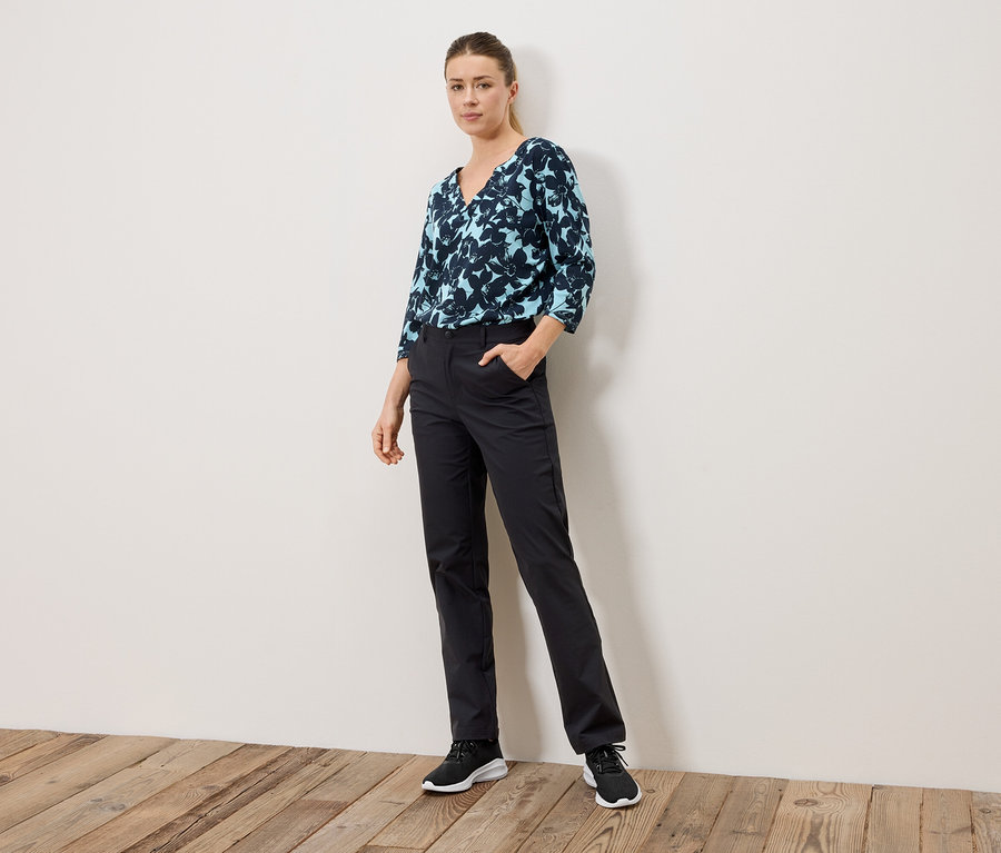 Frau steht mit Hand in der Hosentasche, trägt eine blau gemusterte Bluse und schwarze Hose.